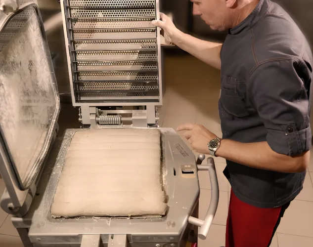 Production-floor image of a baker using the JAC Tradiform hydraulic dough divider and moulder during dough processing and grid-based moulding setup.