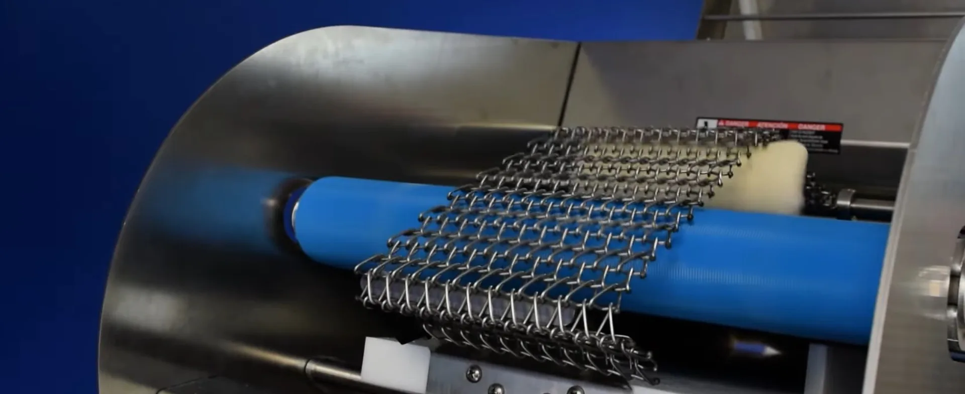 A dough piece moving under a stainless steel curling chain over a blue roller. This is a close-up of the Dough Tech CM275 countertop moulder curling a dough piece during bread and bolillo moulding.