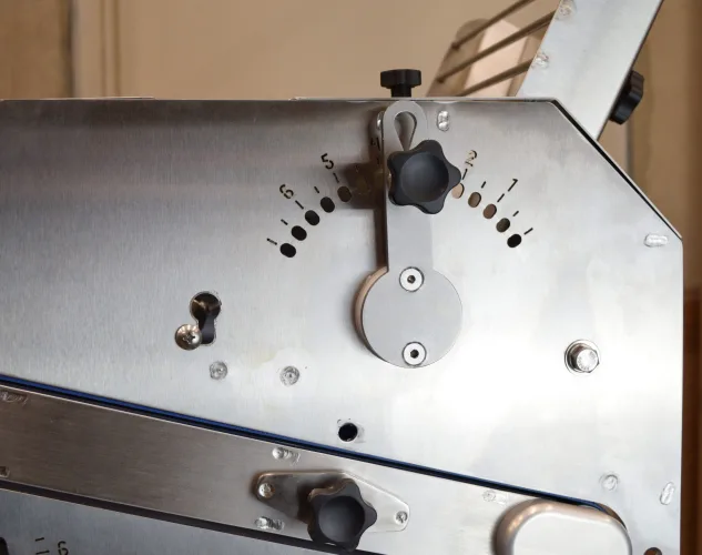 Roller gap adjustment handle with a black locating knob and a numbered hole scale on a stainless steel side panel. This is the roller gap adjustment on the Dough Tech CM275 countertop bread and bolillo moulder.