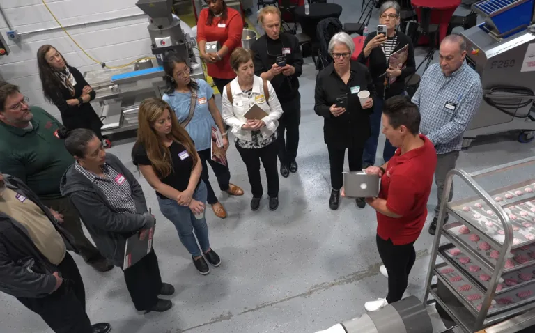 A group of bakers with name tags gathers around an Erika presenter holding a stainless steel container beside a rack of freshly deposited cookies. This live demonstration took place during an open house at Erika's Baker's Lab.