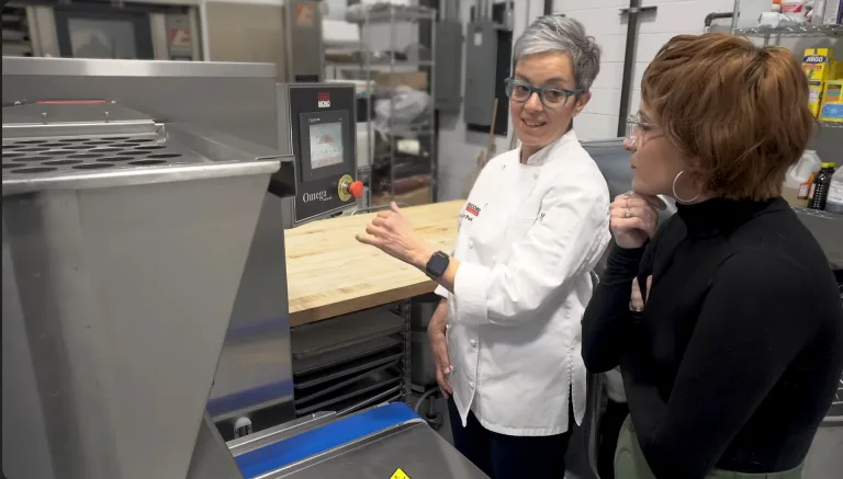 An Erika technical baker in a white chef's coat explains the controls of a MONO Omega depositor to a visitor in a test kitchen. This demonstration took place in Erika's Baker's Lab in West Caldwell, New Jersey.